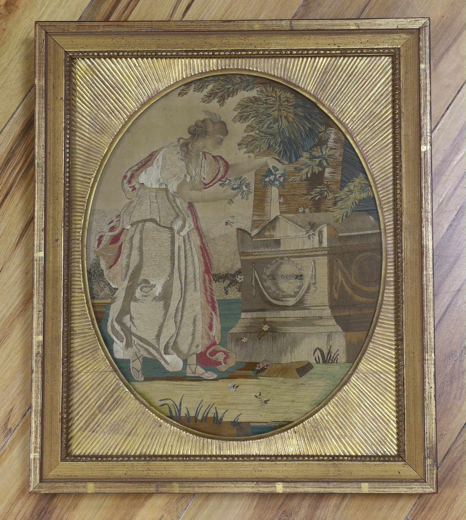 A gilt framed Regency oval silkwork picture of a classical maiden scattering flowers at a tomb, labelled Cowper, probably commemorating the death of the poet William Cowper in 1800, 30x25cm